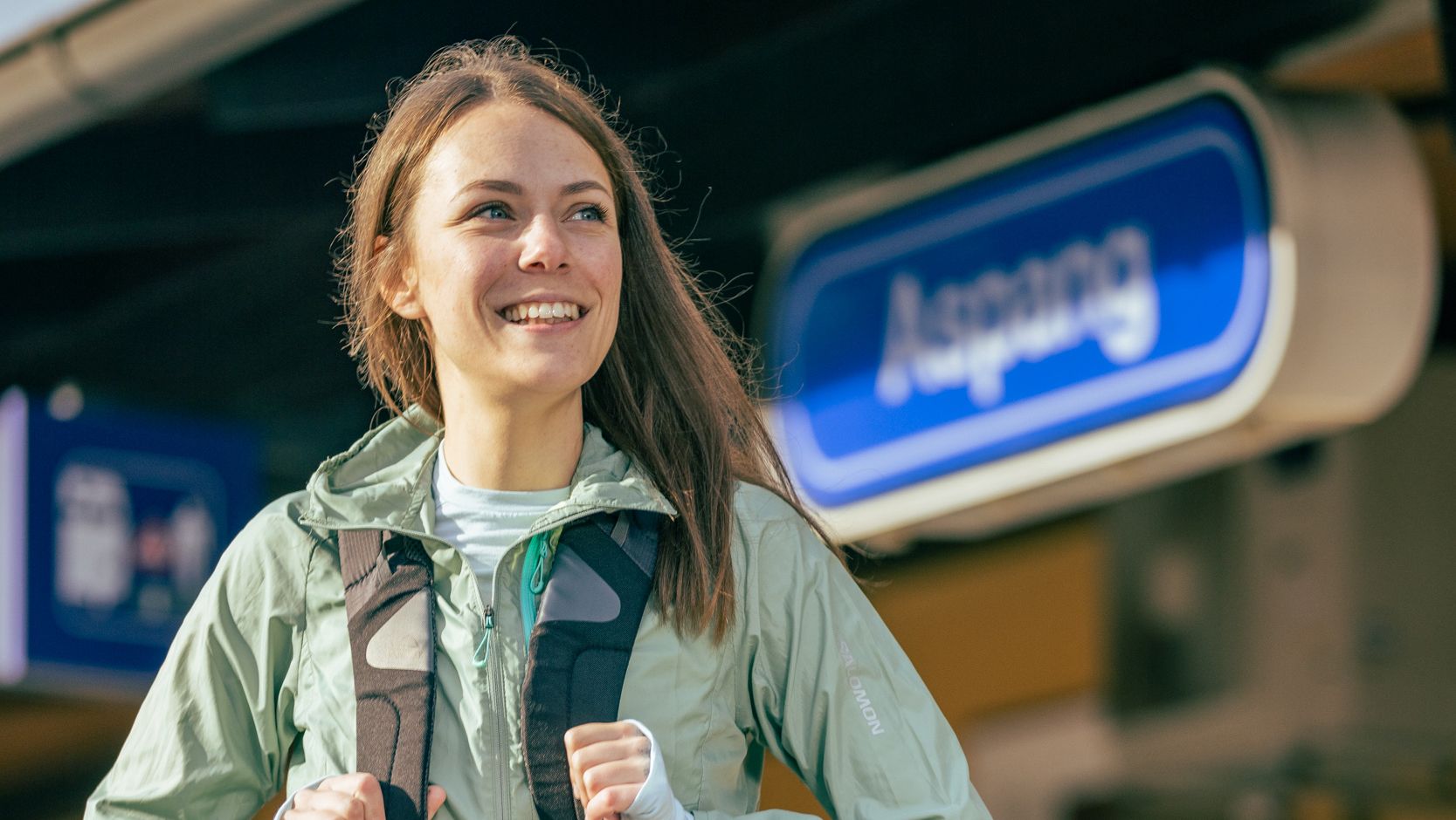 A smiling woman with a rucksack stands in front of a station sign that reads 'Aspang'.