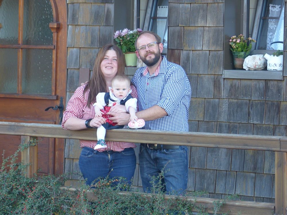 A couple with a baby stands in front of a wooden house.