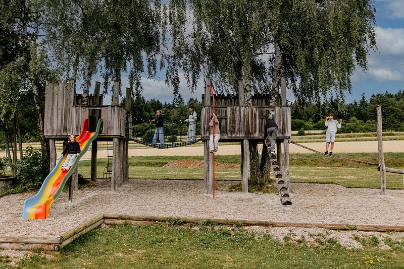 Children play on a wooden outdoor playground with a slide and rope bridge.