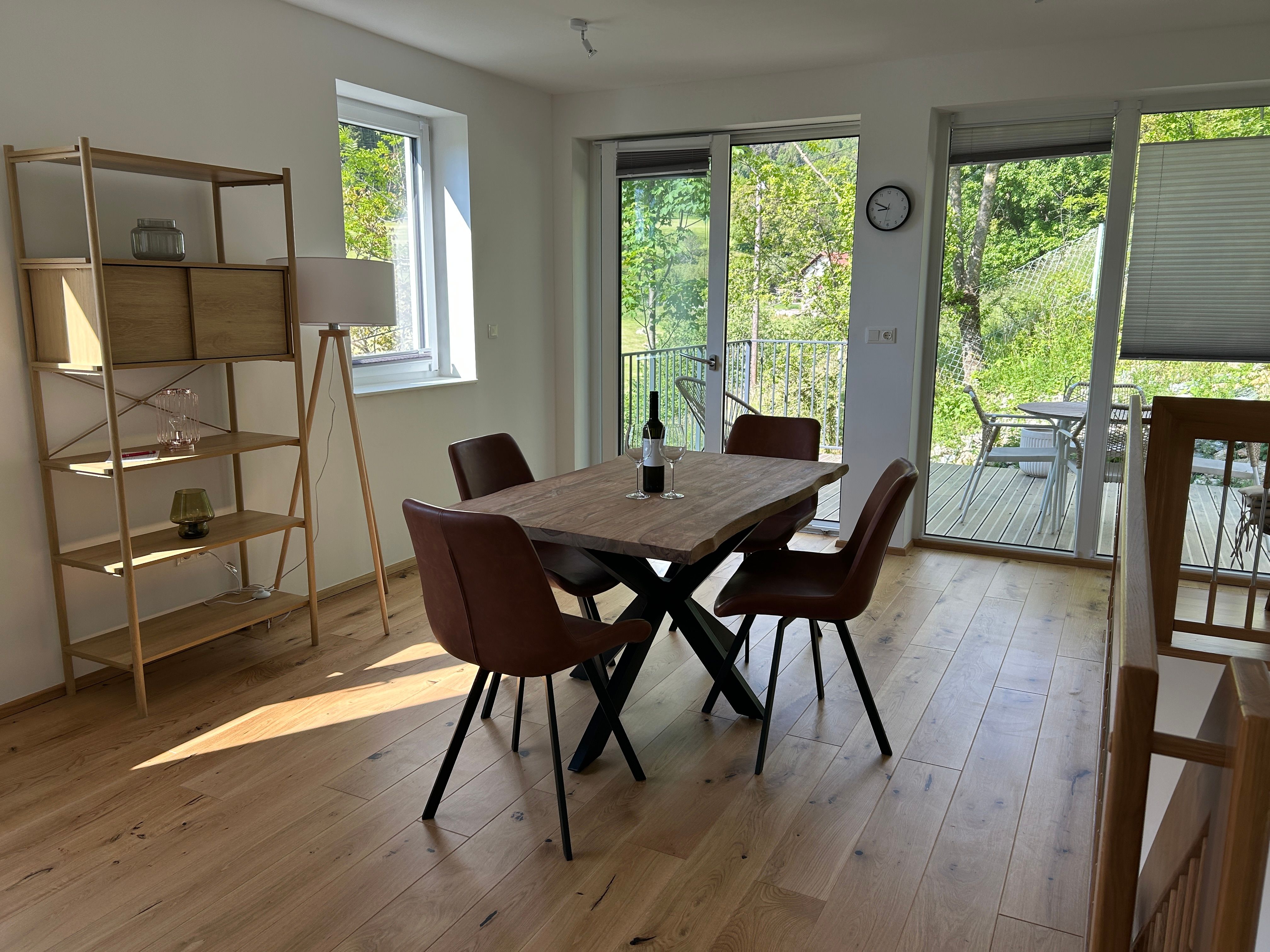 Interior view of a modern living room with wooden floor, dining table and chairs, shelf and view of a terrace with garden.