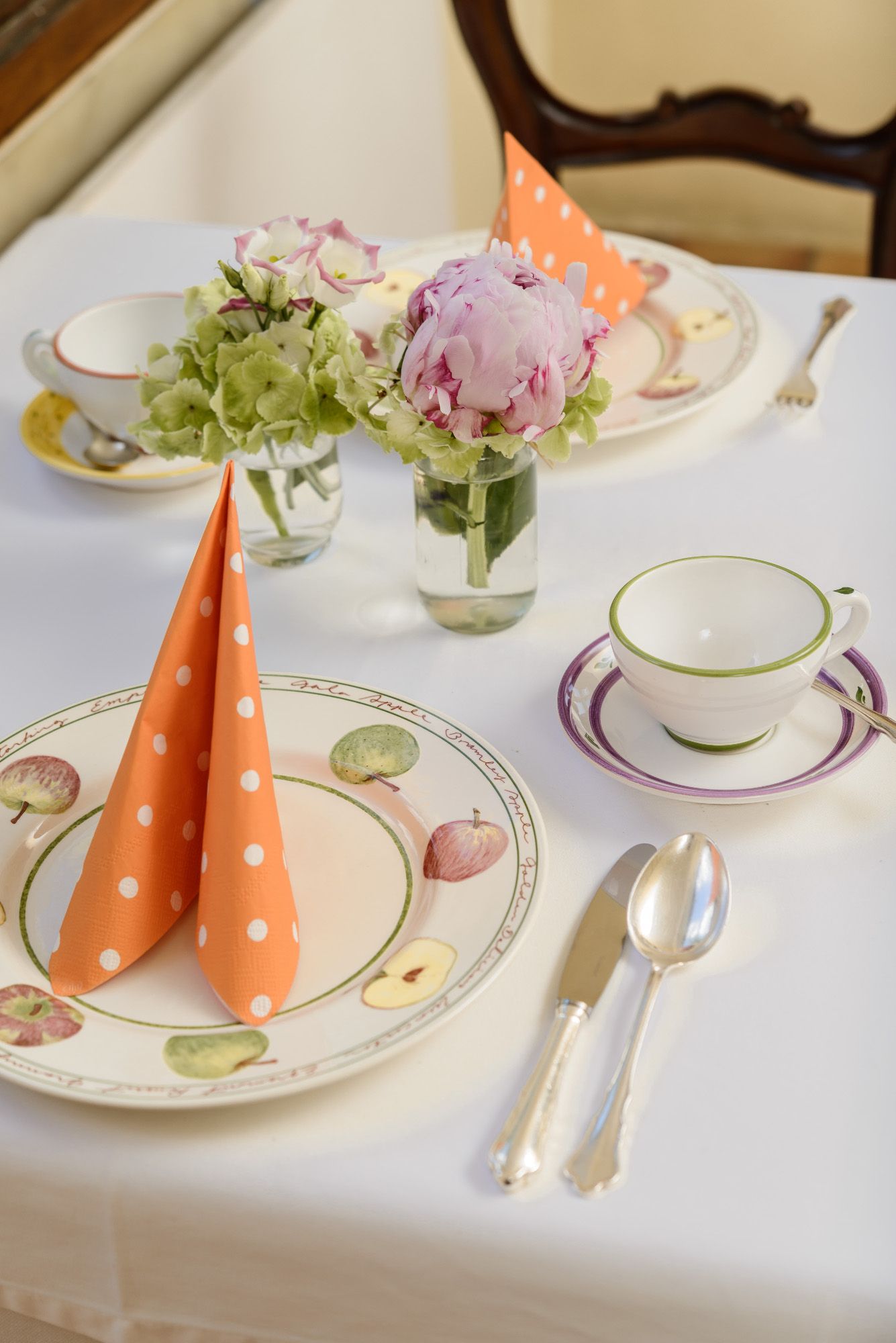 An elegantly laid table with flowers, orange dotted napkins and crockery with fruit motifs.