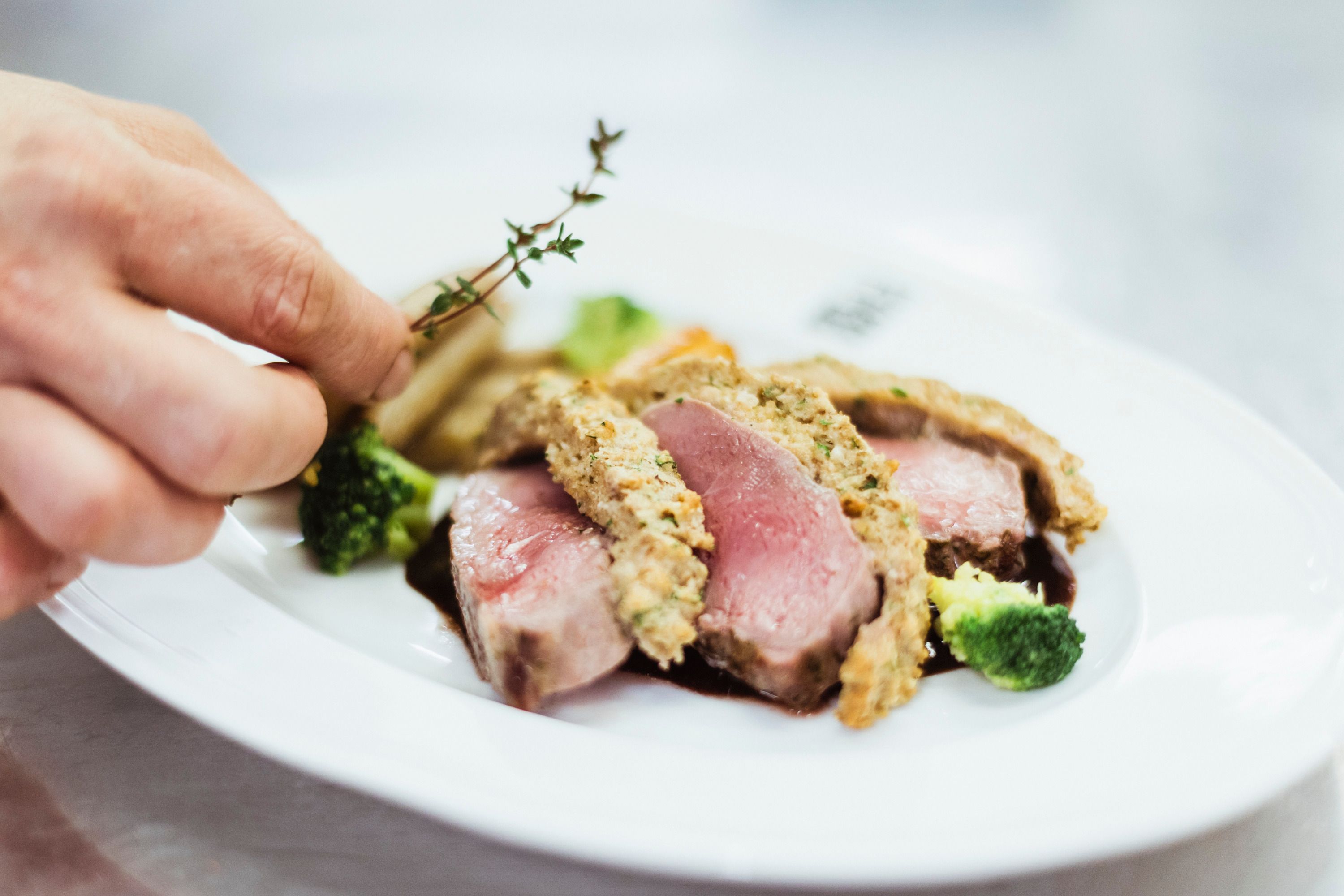 Close-up of a pink roasted meat dish with herb crust, broccoli and a hand adding thyme.