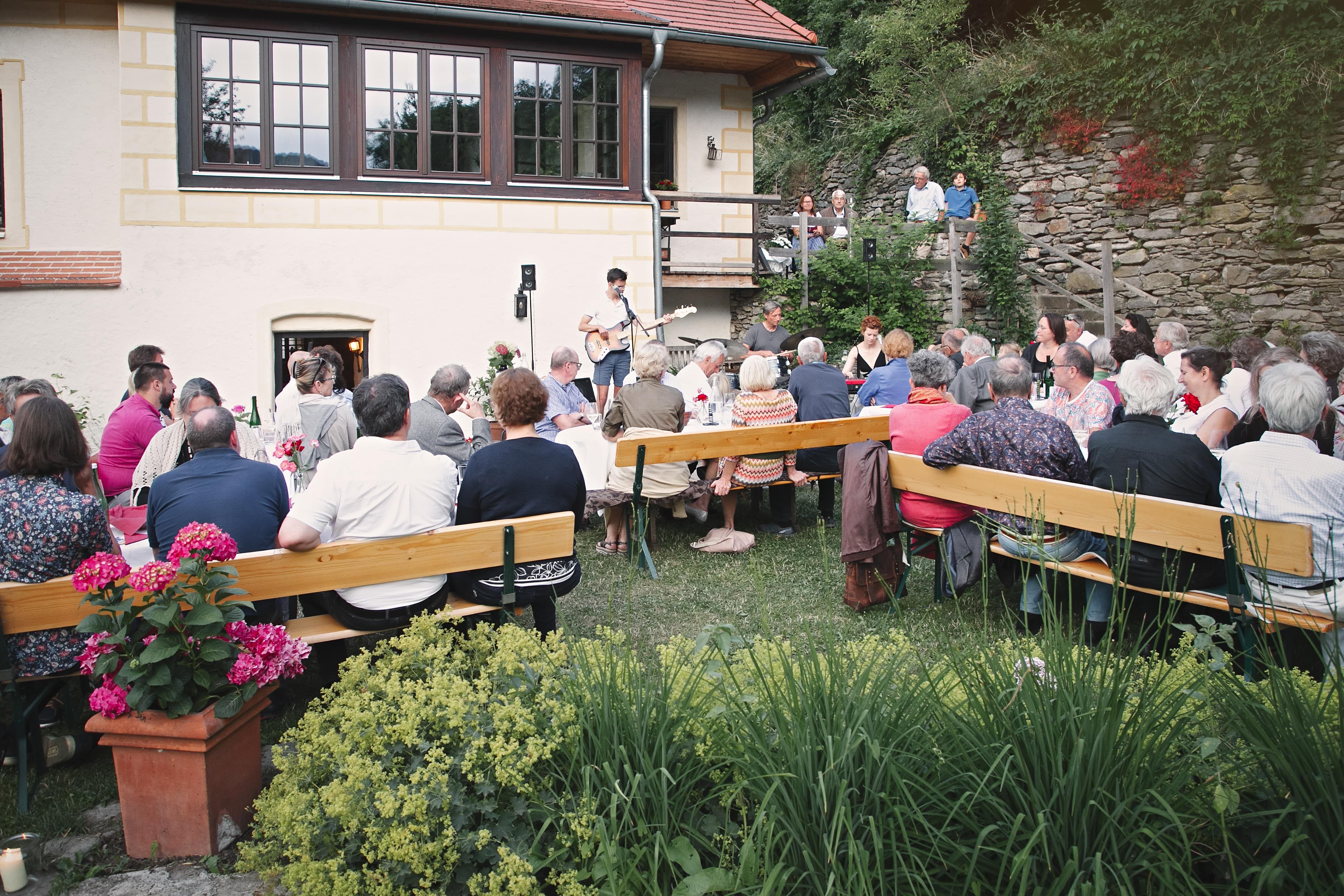 People sit outside at long tables at a wine tavern while a musician performs.