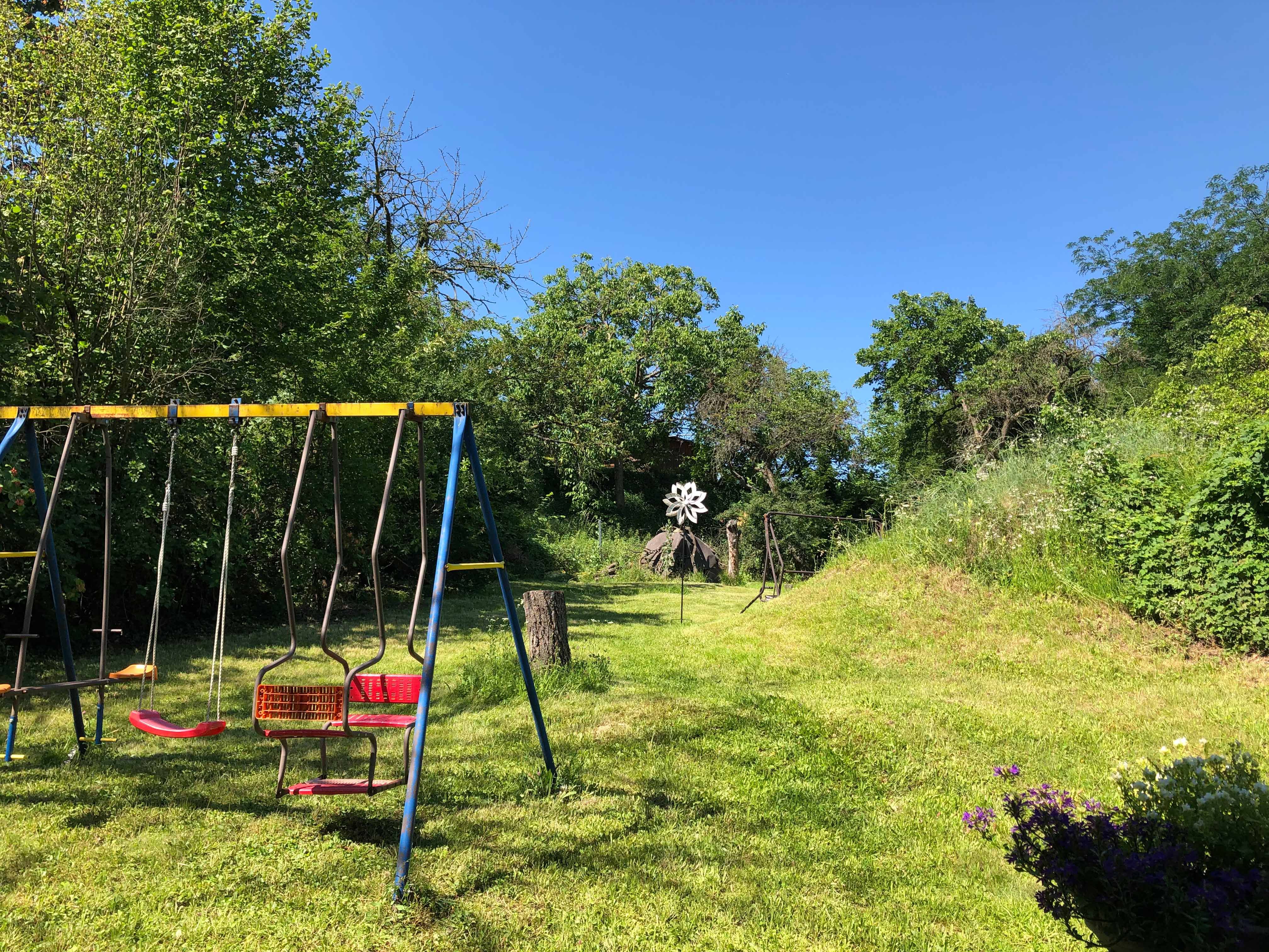 A garden with a swing, surrounded by trees and meadows, under a blue sky.
