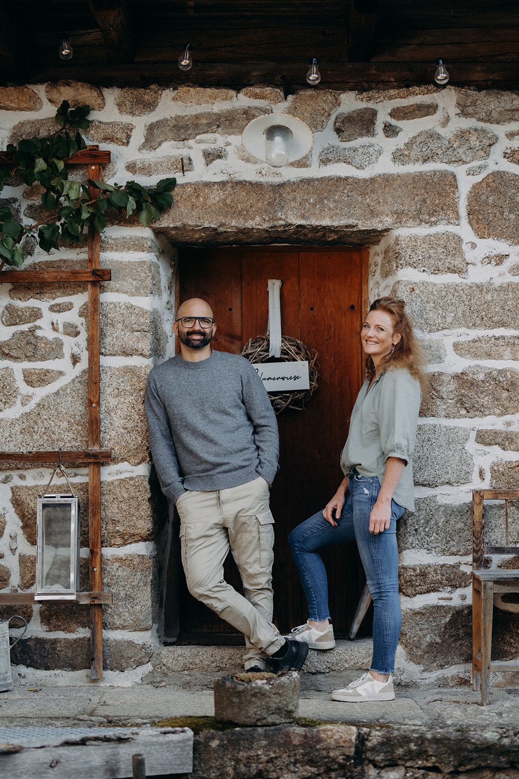 Two people are standing in front of a rustic stone house wall with a wooden door.