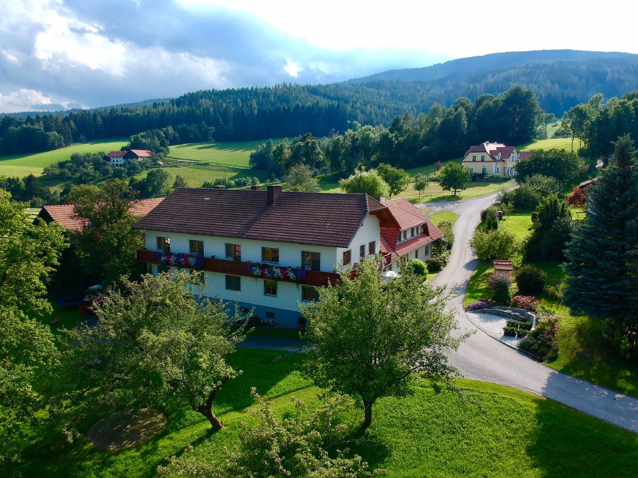 Rural landscape with a large house, surrounded by trees and meadows, under a cloudy sky.
