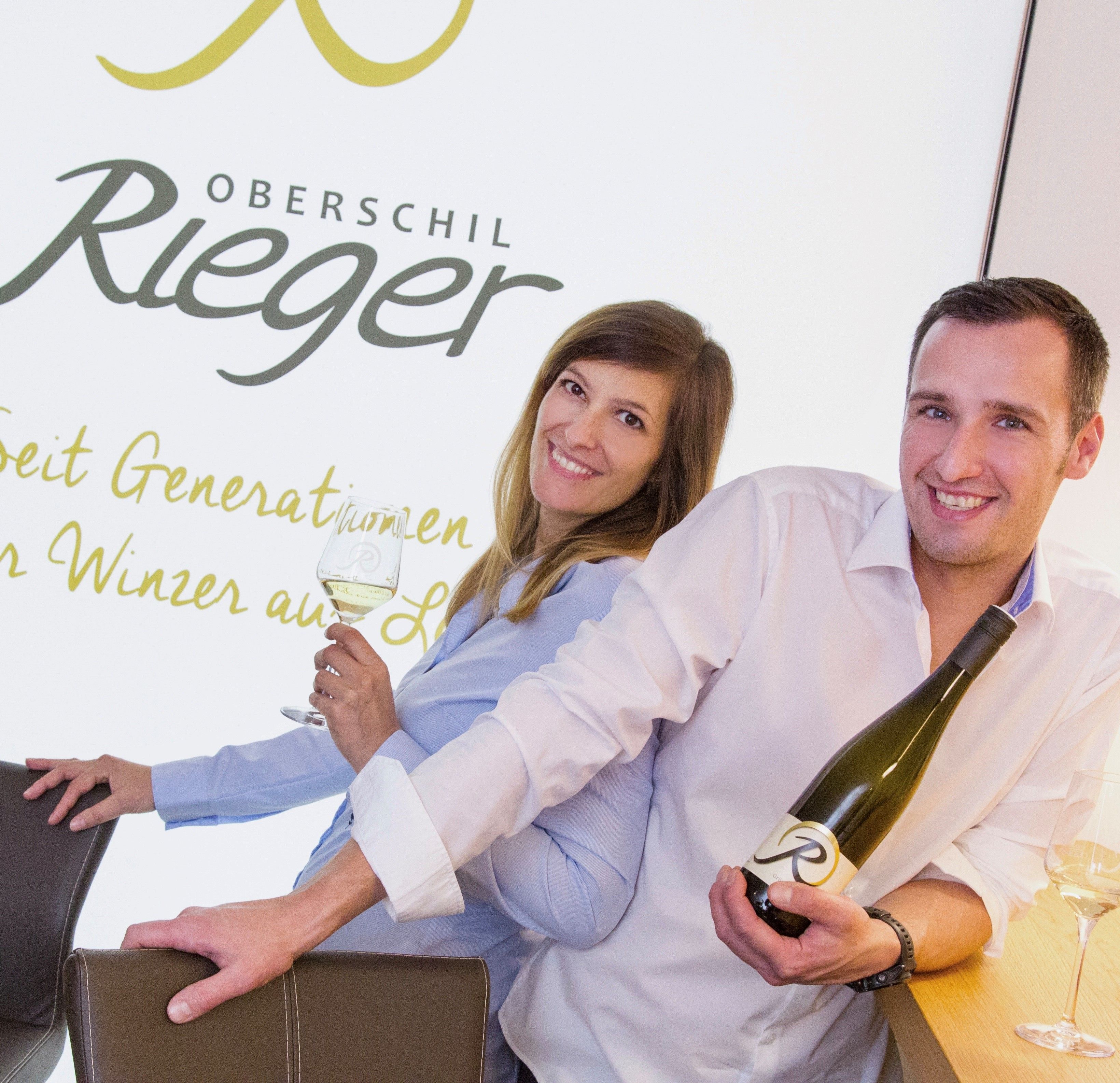 Two people are smiling and holding bottles of wine in front of a sign that reads 'Oberschil Rieger'.