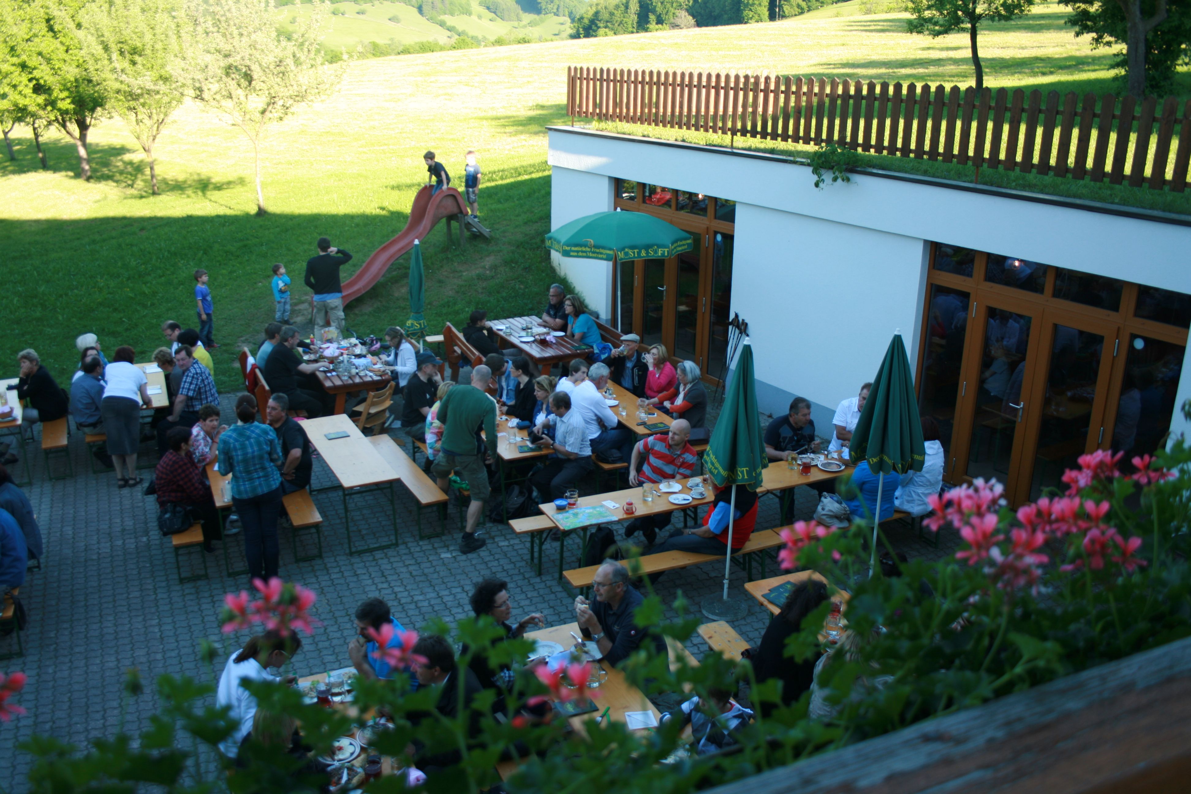 People sit at tables outside, surrounded by greenery and flowers.