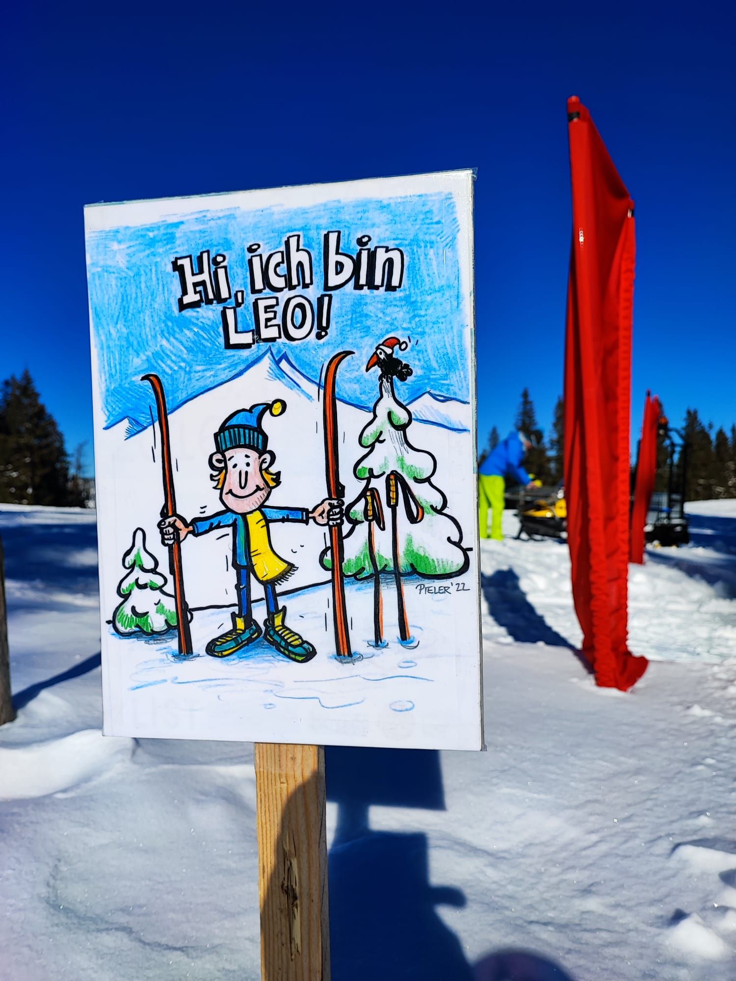 A sign in the snow with a drawing of Leo, a cartoon character with skis, against a blue sky.