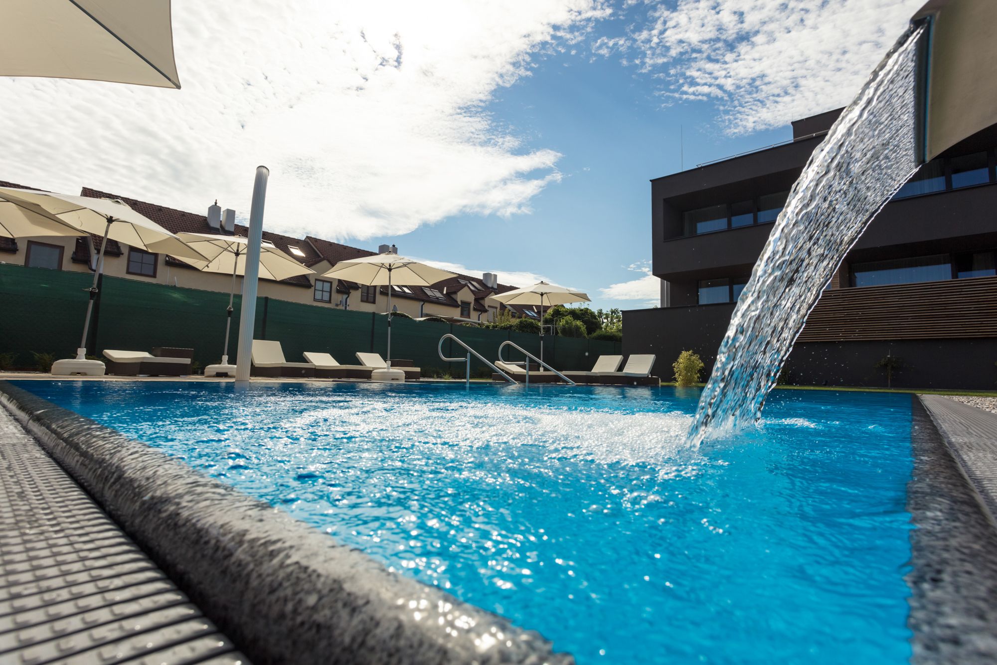 A modern pool with waterfall, surrounded by sun loungers and parasols, in sunny weather.