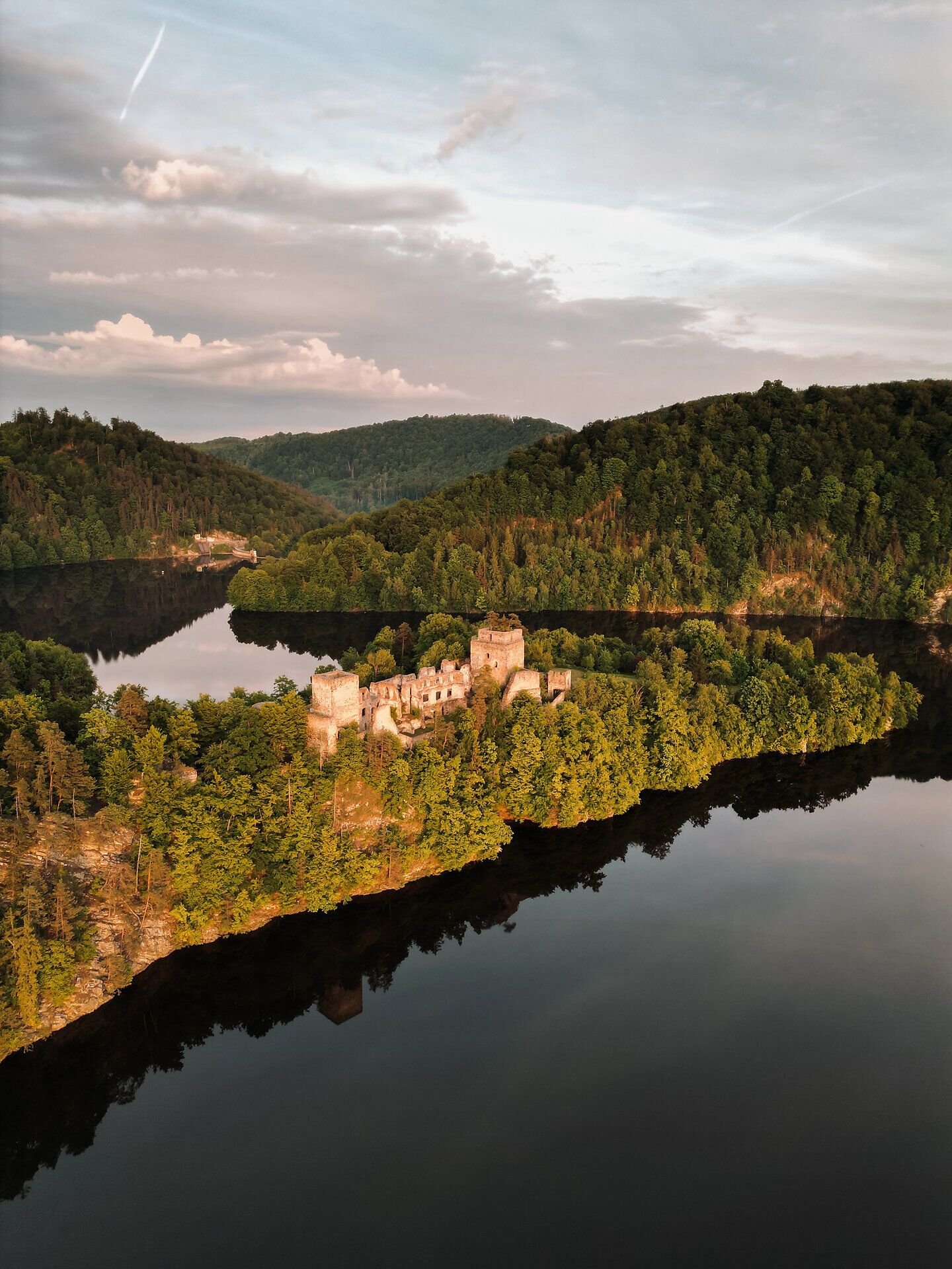 Aerial view of a castle ruin by the lake, surrounded by forest and hills.
