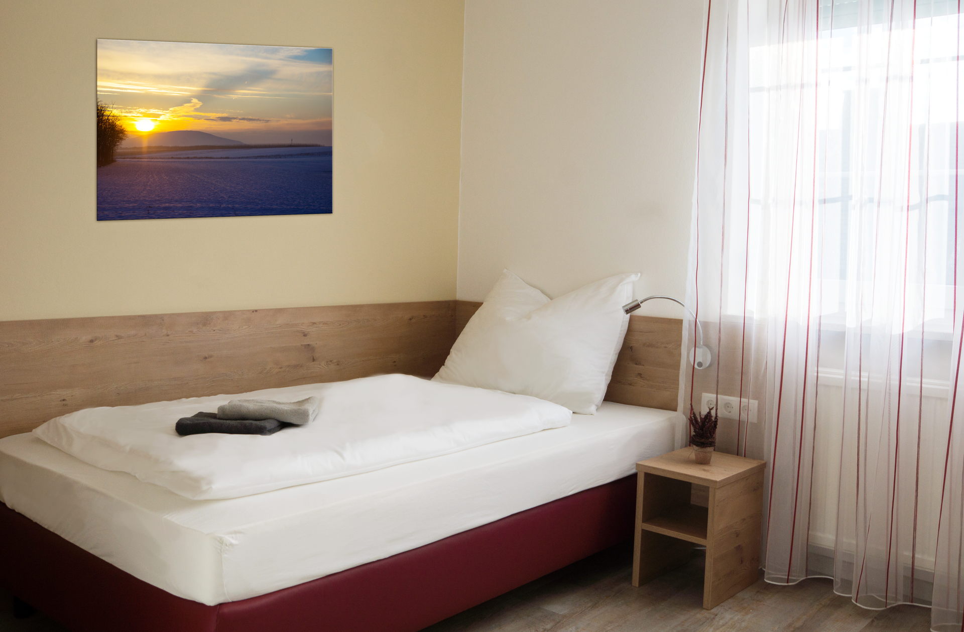 A simple single room with a bed, a bedside table and a picture on the wall. A window with curtains lets in light.