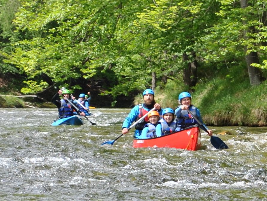 People in canoes paddling on a river in a green forest landscape.