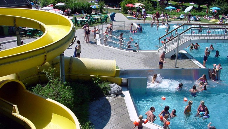 An outdoor pool with a yellow water slide, swimmers in the pool and people on the sunbathing lawn.