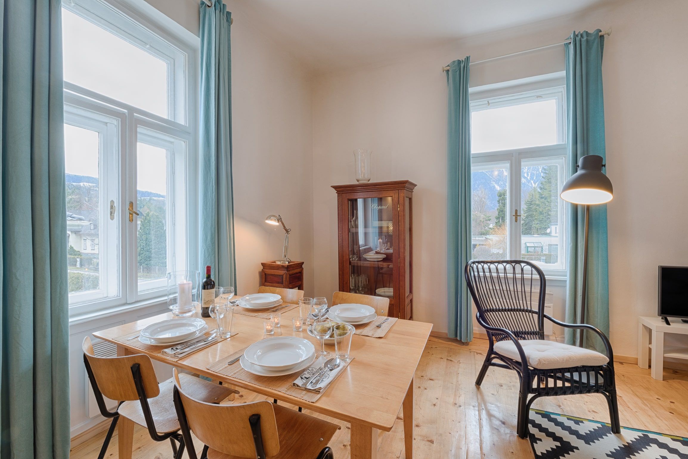 Dining area in a living room with wooden table, chairs, display cabinet and large windows with blue curtains.