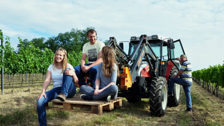 Four people in a vineyard next to a tractor, three sitting on a pallet, one leaning against the tractor.