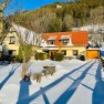 Snow-covered house with red roof and garden, surrounded by trees and fence, in sunny weather.