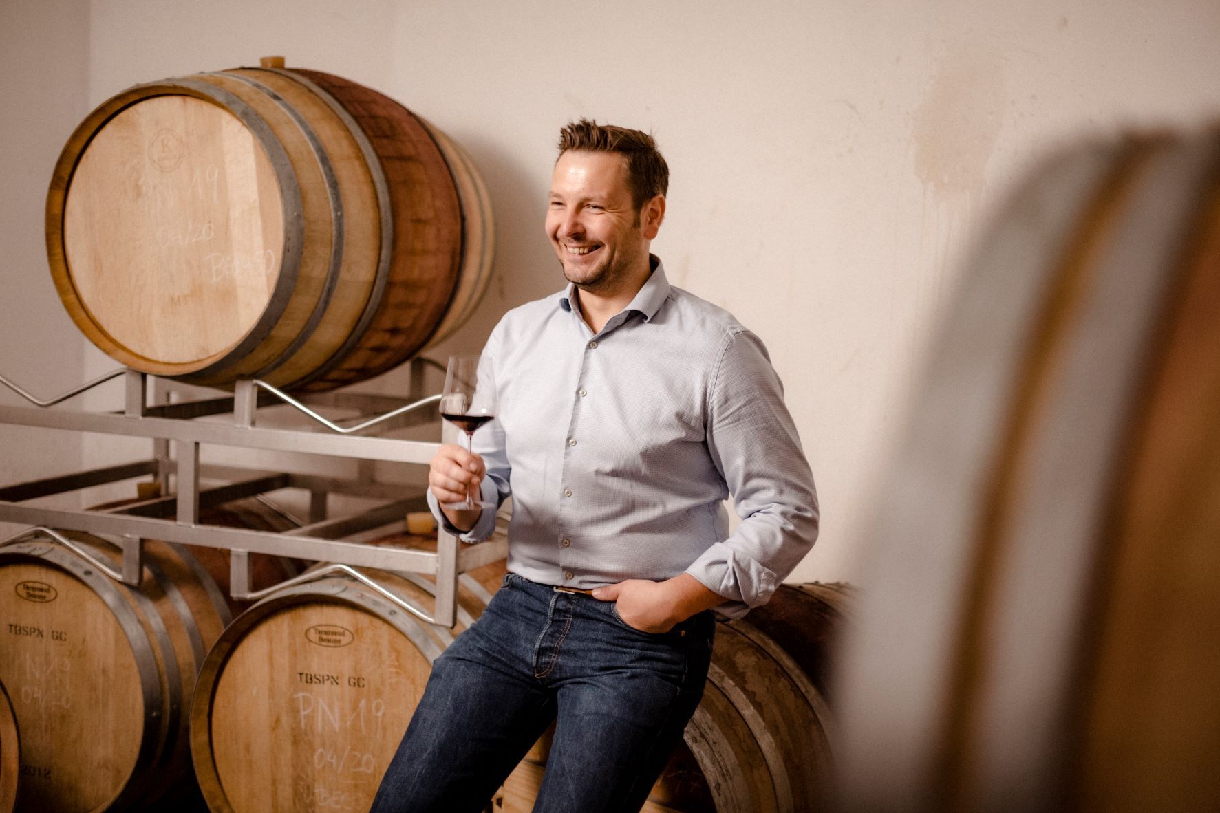 A man leans smiling against wine barrels and holds a glass of red wine in his hand.