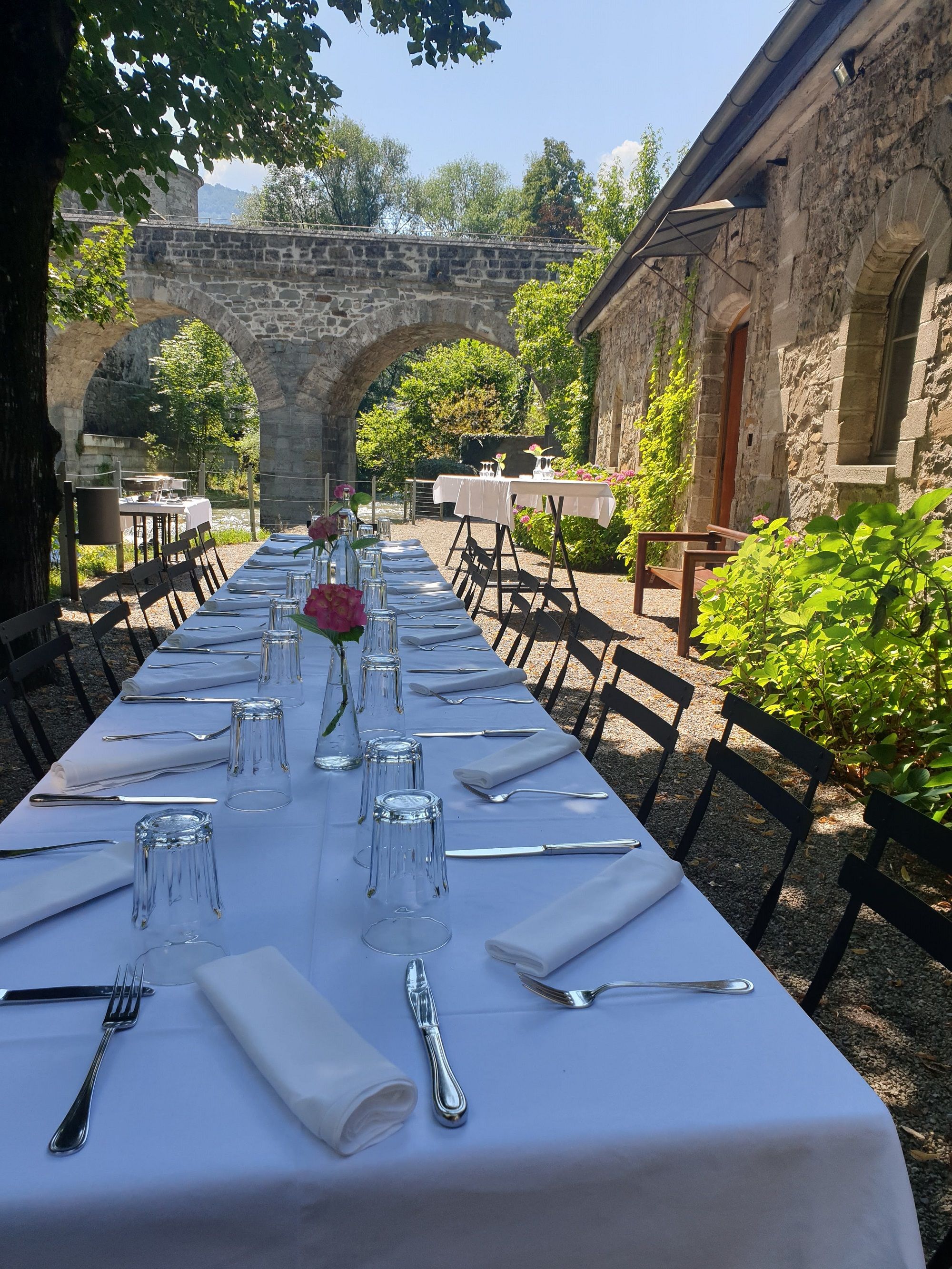 Long outdoor table with white tablecloth, set with glasses and cutlery, stone arches and green vegetation in the background.