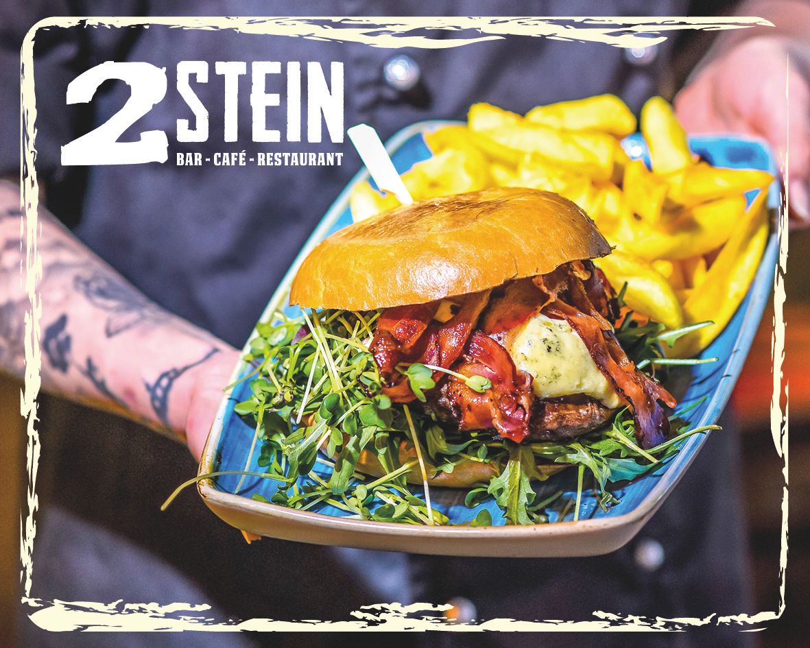 A burger with bacon, cheese and rocket on a plate with fries, presented by a tattooed hand. Logo of 2Stein Bar-Café-Restaurant top left.