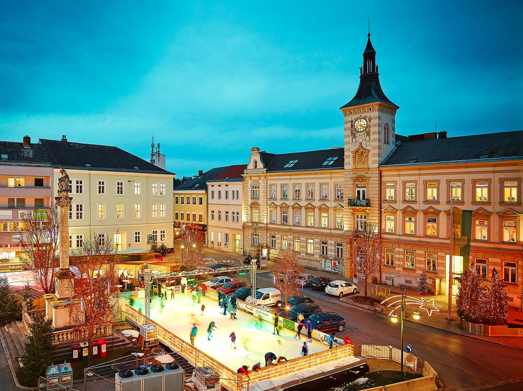 Ice rink in a town at dusk, surrounded by historic buildings and Christmas lights.