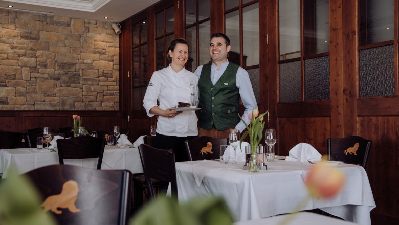 A female chef and a man in traditional dress stand smiling in an elegant restaurant.
