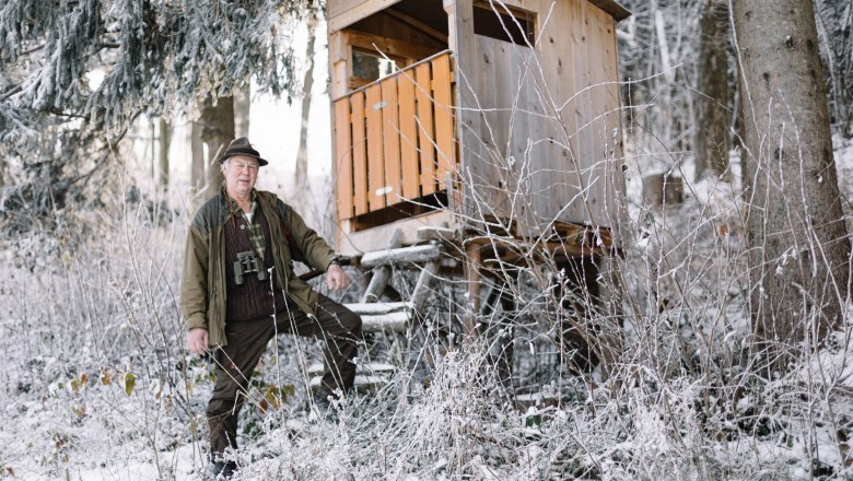 A man in hunting clothes stands next to a high seat in the snowy forest.