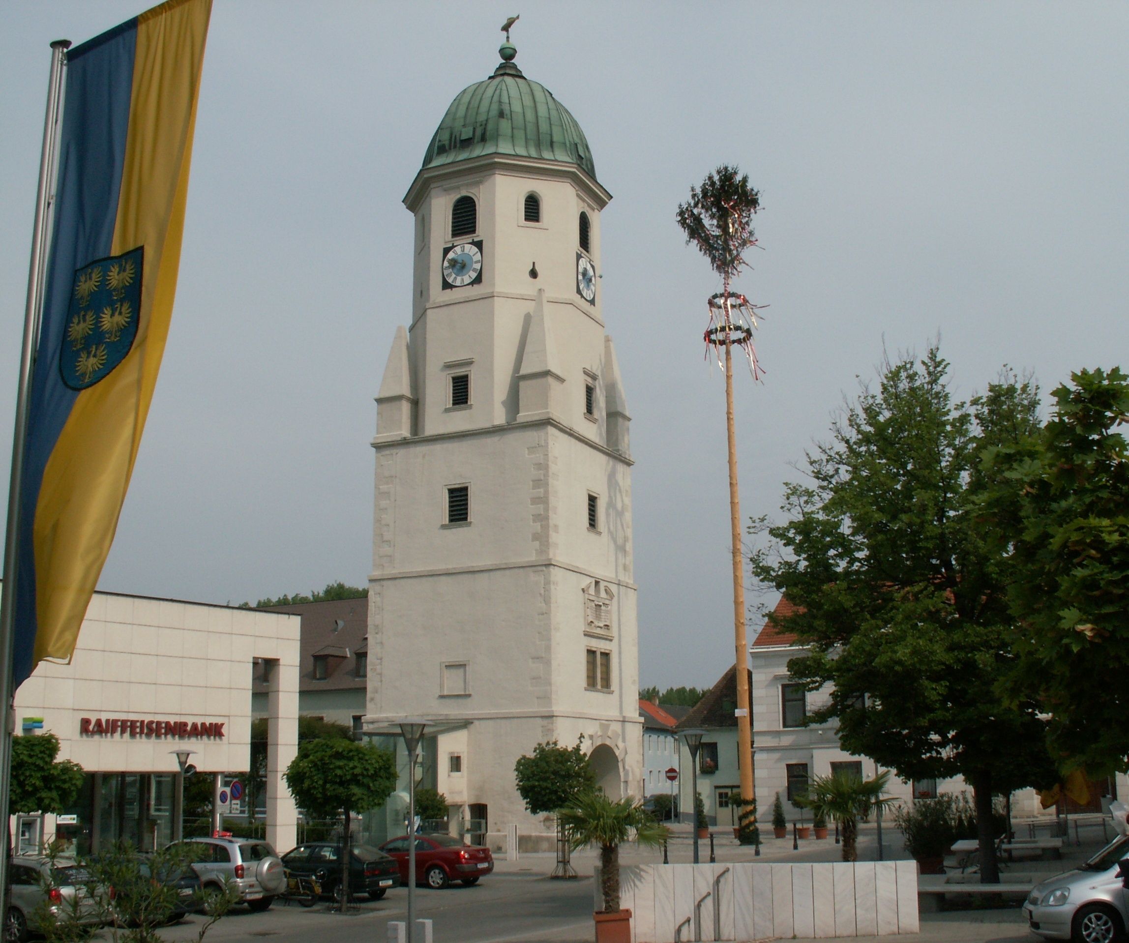 A church tower with a clock and green roof in Fischamend, next to a maypole and a Raiffeisenbank branch.
