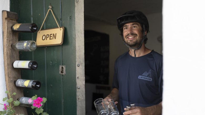A man with a bicycle helmet stands smiling in an open doorway, holding glasses and bottles. An "Open" sign hangs on the door.