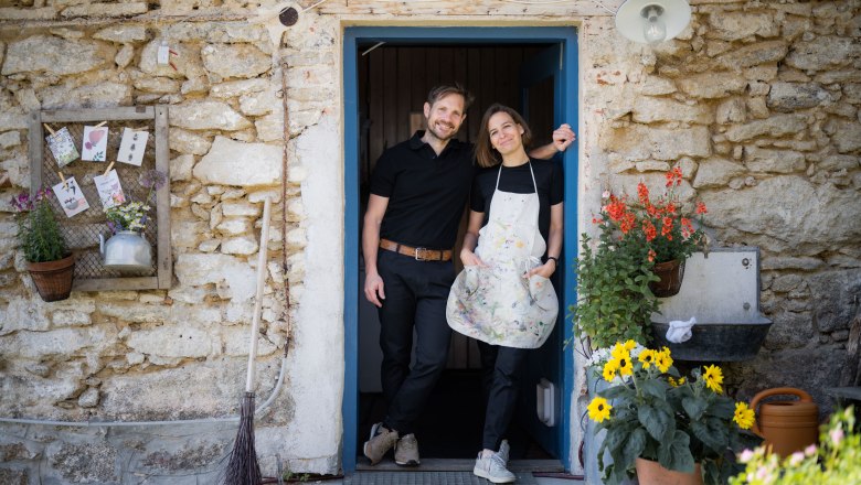 A man and a woman stand smiling in a doorway of a rustic stone house, surrounded by flowers and decorations.