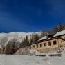 A wooden house in the snow against a mountain backdrop under a clear blue sky.