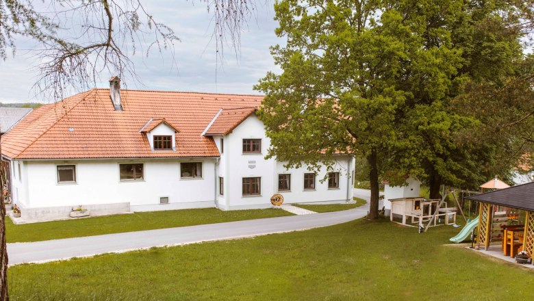 Knappenhof, © Katja Koppensteiner Fotografie A white building with a red tiled roof, surrounded by trees and a meadow, with a playground in the foreground.
