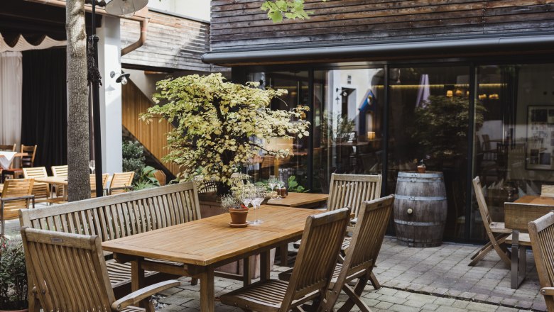 A cozy courtyard with wooden furniture, plants and a wine barrel, surrounded by buildings with wooden cladding.