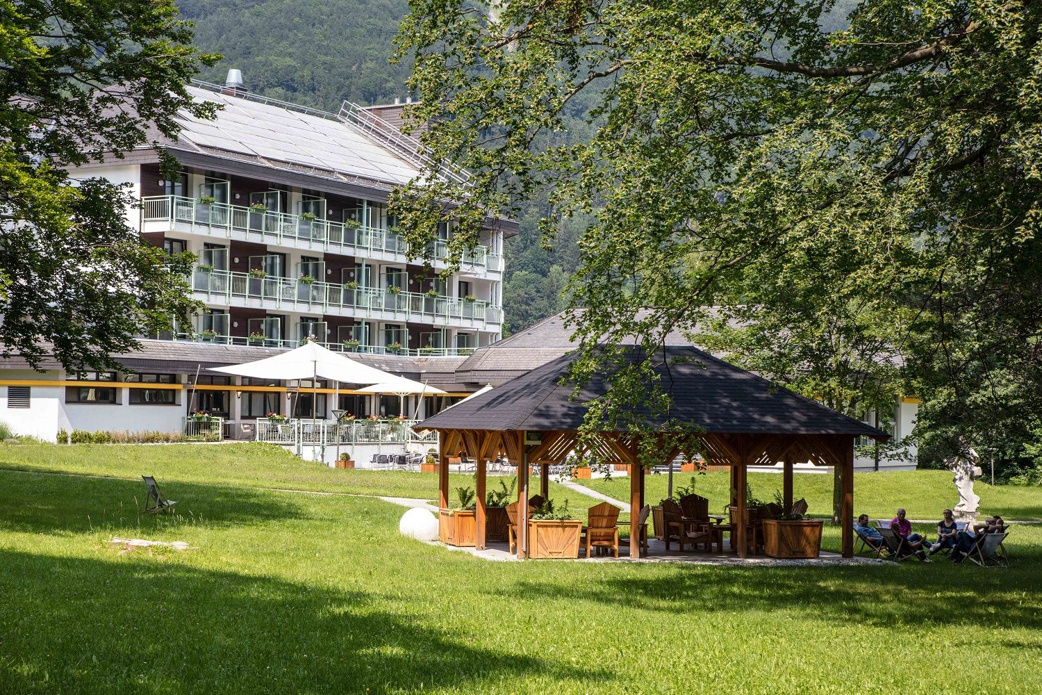 A hotel building with balconies in the background, a pavilion and people on chairs in the foreground, surrounded by trees and lawn.
