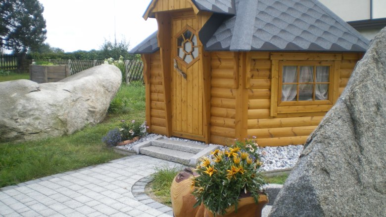 A small, wooden barbecue hut with a gray roof, surrounded by stones and plants.