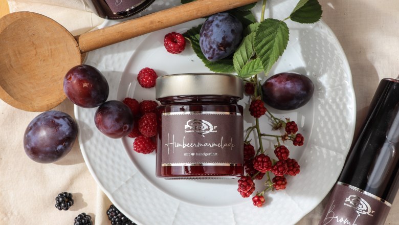 A jar of raspberry jam on a plate with fresh plums, raspberries and blackberries, surrounded by a wooden spoon and a bottle of blackberry syrup.