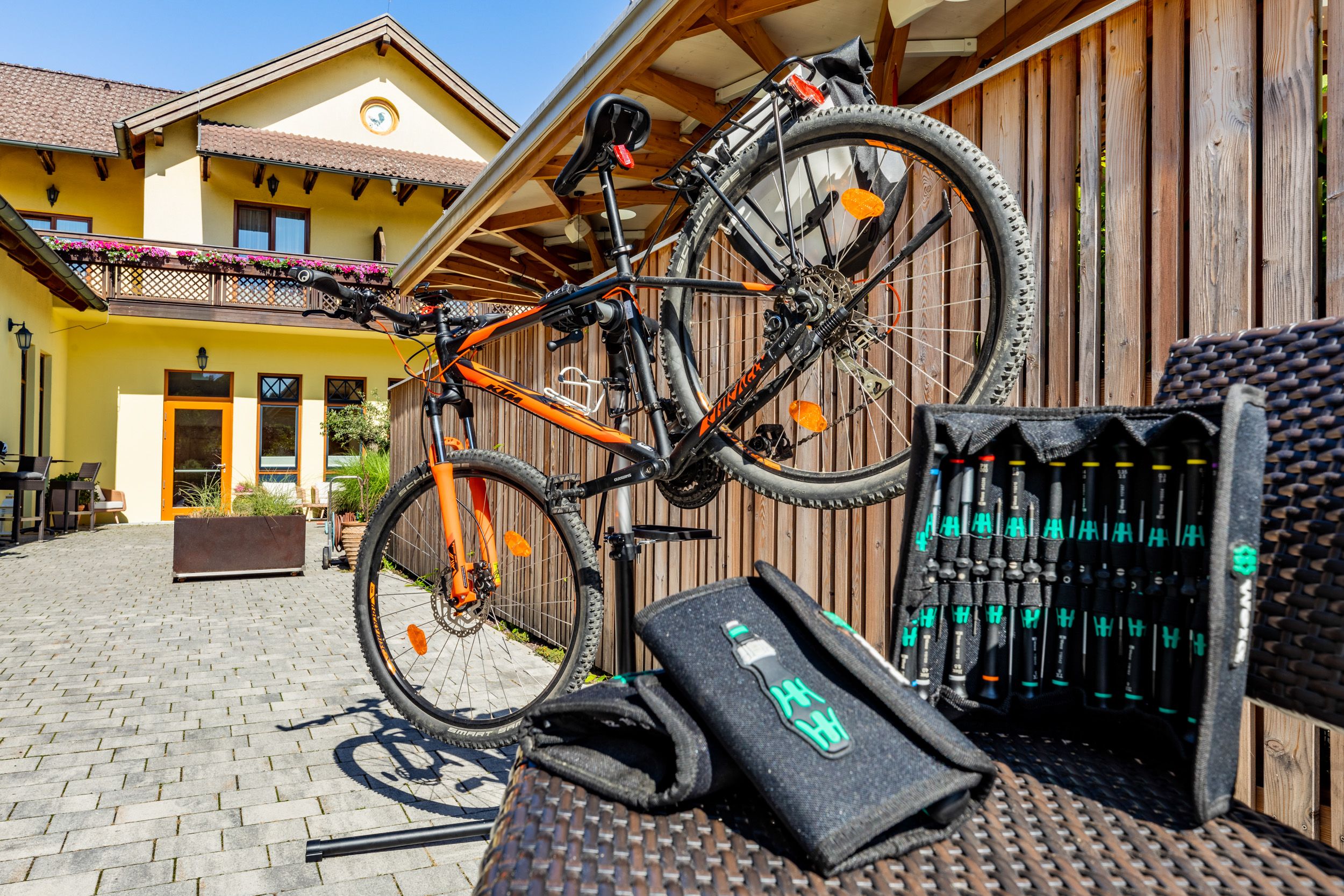 An orange bicycle on a stand in front of a yellow building with a wooden fence and tool set in the foreground.