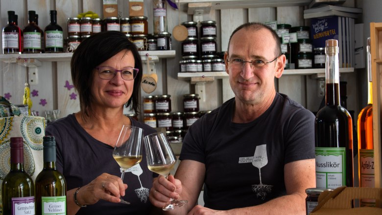 A man and a woman sit at a table in a farm store, holding glasses of wine and smiling. Shelves with products can be seen in the background.