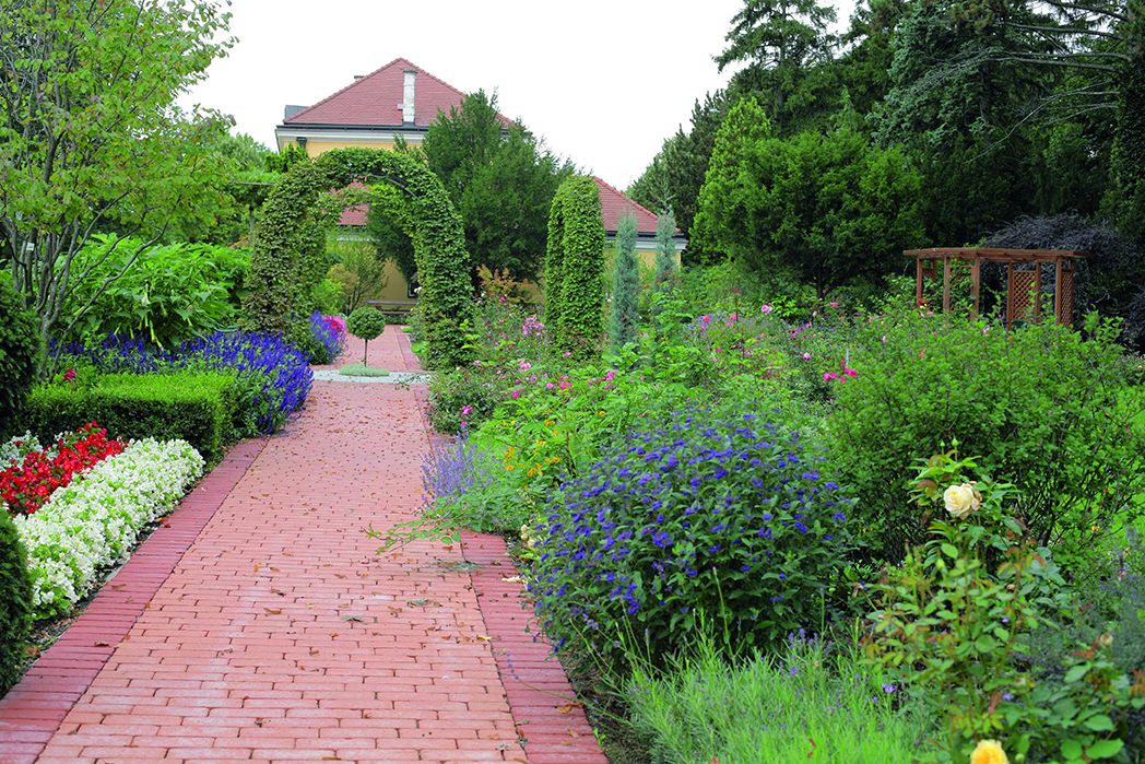 A well-kept garden with a red brick path, surrounded by colorful flowers and green bushes, leads to a house with a red roof.