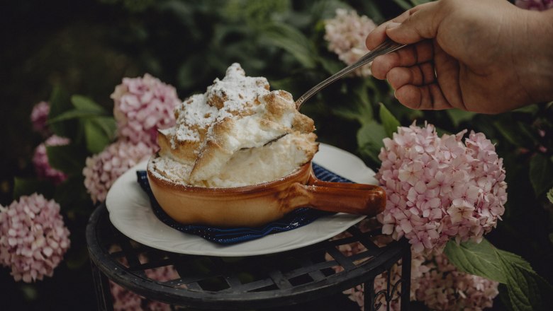 A soufflé in a brown bowl, surrounded by pink hydrangeas, is served with a spoon.