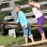 Two children balance on a wooden raft over water, in the background a wooden bridge.