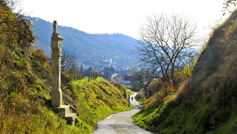 A narrow path leads through a hilly landscape with a stone monument and bare trees in the Kamp Valley ("Kamptal" in German) Schönberg Nature Park.