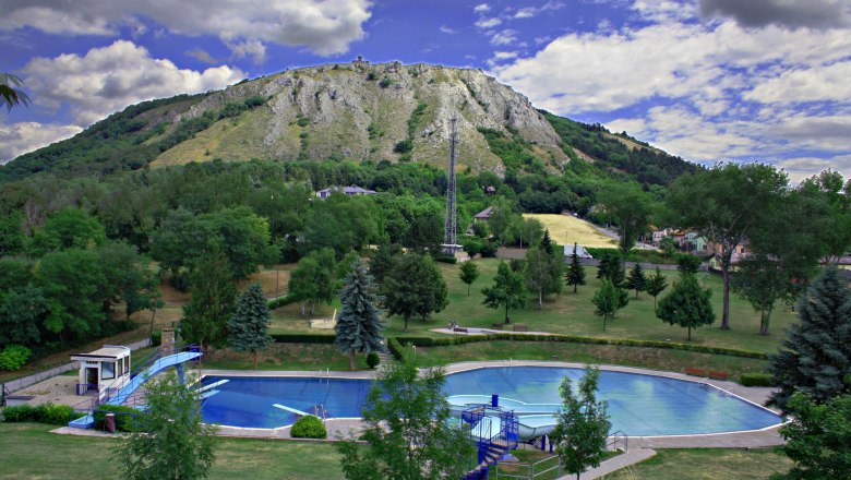 An outdoor pool with a round pool and a slide, surrounded by trees, in front of a wooded hill with a rocky peak.