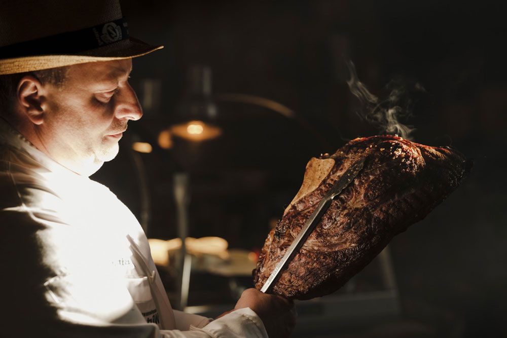 A man in a white shirt holds a large, grilled piece of meat with a knife.