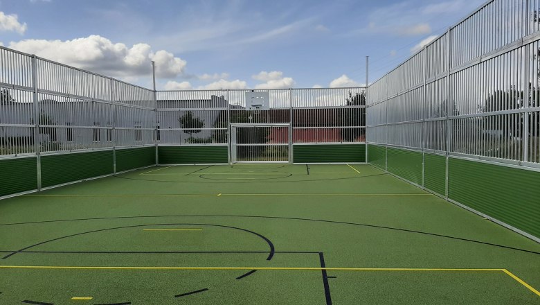 An empty, fenced multi-sports field with a green floor and markings for various sports under a blue sky with clouds.