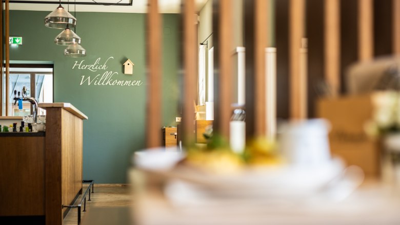 Interior view of a modern guest room with a green wall and "Welcome" lettering. Lamps hang above the wooden bar and a small birdhouse decorates the wall.