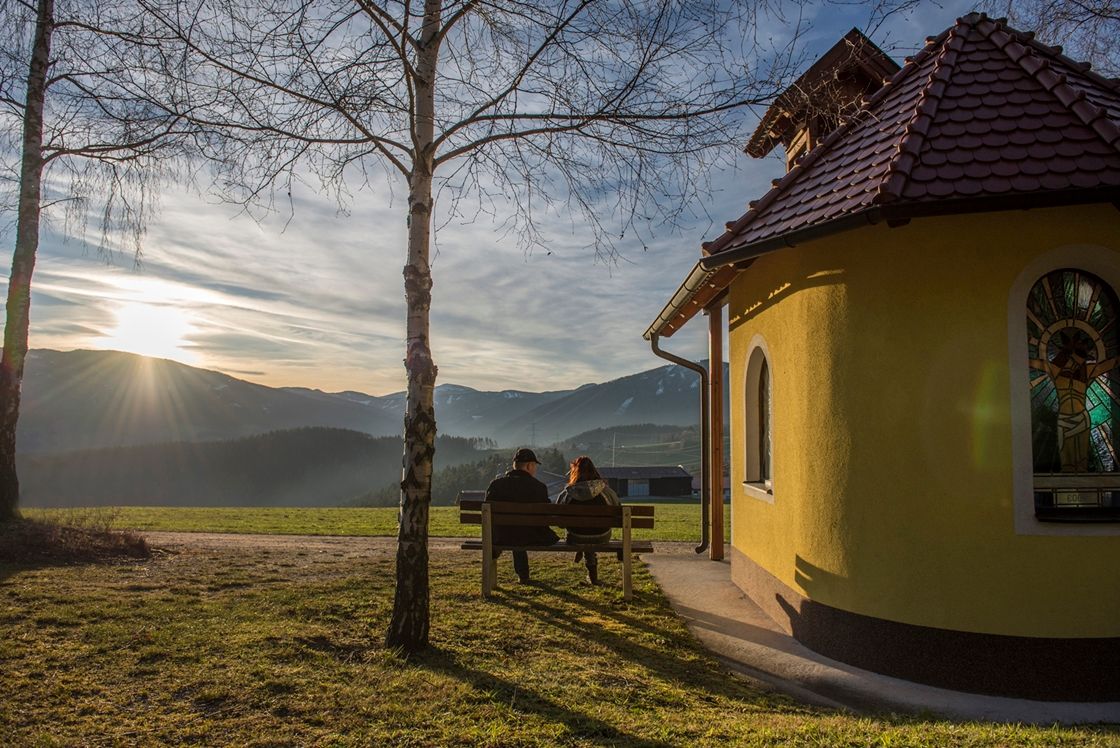 Two people sit on a bench next to a small chapel with a view of the mountains at sunset.