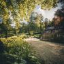 A rural garden with a small house and a gravel path, surrounded by trees and plants in the sunlight.