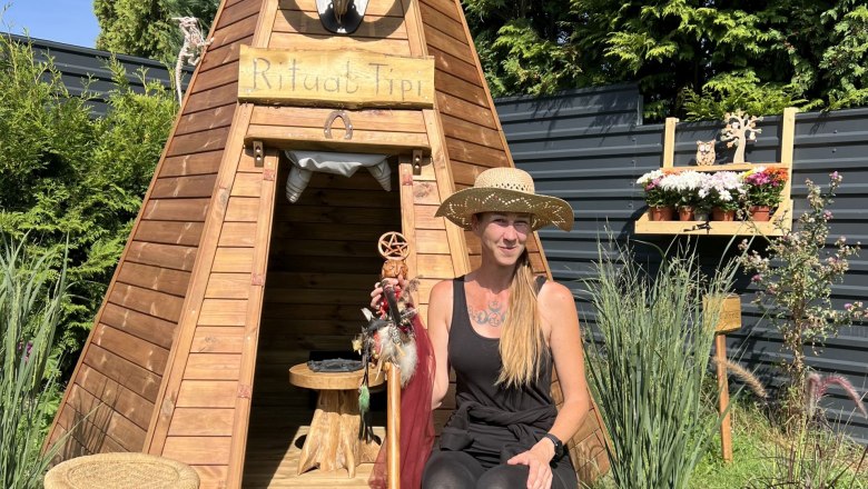 Woman in a straw hat sits in front of a wooden tepee in the garden.