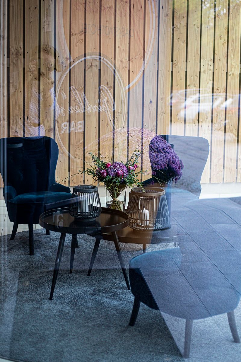 A cozy lounge area with blue armchairs, a table with candles and a bouquet of flowers, reflected in a glass wall.